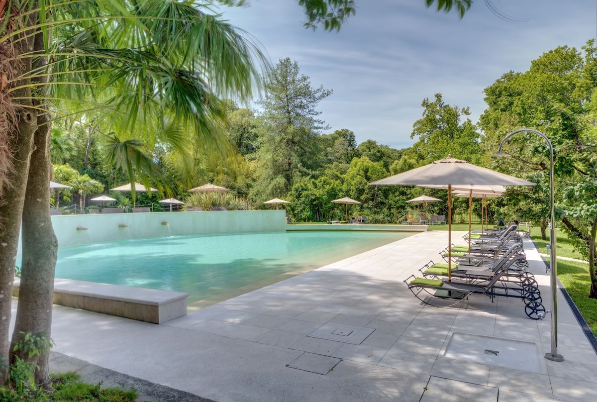 A serene pool area is surrounded by lush greenery, with lounge chairs and umbrellas positioned along the edge. The tranquil water reflects the sky and is complemented by the landscaped garden that provides a calming atmosphere.