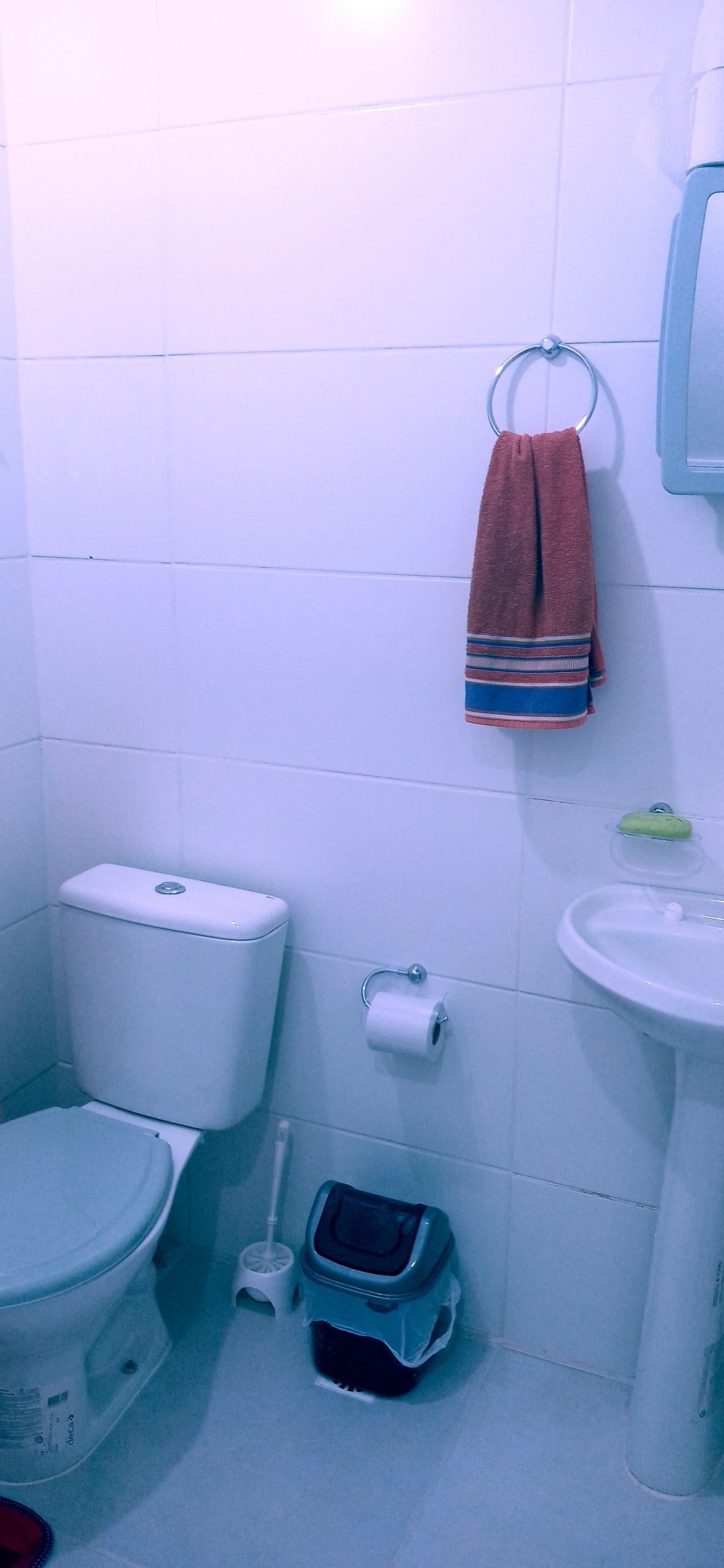 A clean bathroom is presented, featuring white tiled walls and a compact layout. A toilet and a small sink with a mirror are visible. A towel hangs from a ring beside the sink, and a toilet brush holder is placed on the floor.