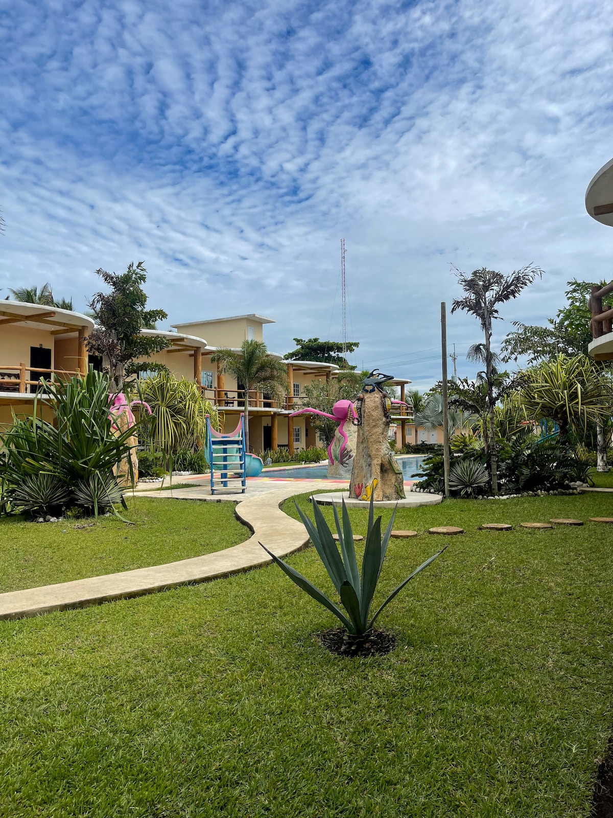 A well-maintained garden features winding stone pathways and lush greenery surrounding a pool area. Vibrant plants and a decorative stone sculpture add character. Two-story buildings with separate balconies are visible in the background, creating a welcoming outdoor environment.