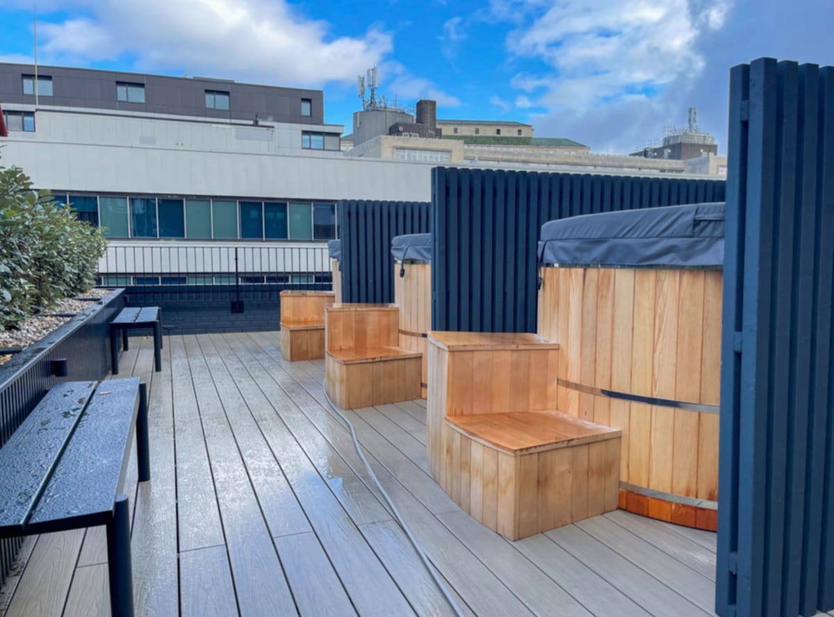 A modern rooftop space features several wooden hot tubs arranged on a wooden deck, complemented by simple, sturdy wooden seating. The surroundings include low, dark wood partitions and a backdrop of city buildings under a bright sky.
