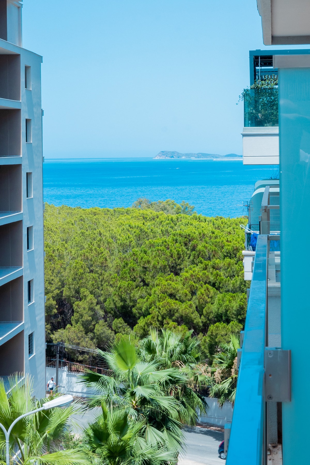 A view from the balcony showcases clear blue waters and distant islands framed by lush greenery. Palm trees line the foreground, creating a natural contrast against the vibrant sea. Nearby buildings reflect the sunlight, enhancing the bright coastal atmosphere.