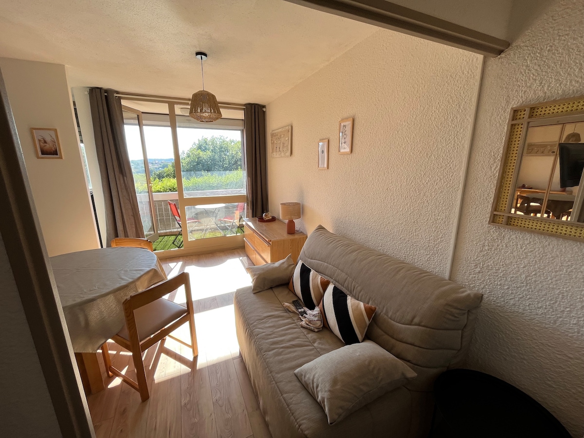 A light-filled living area displays a comfortable sofa adorned with decorative cushions. A small dining table can be seen beside a wooden dresser. Large glass doors open to a balcony with outdoor seating, offering views of the surrounding greenery.