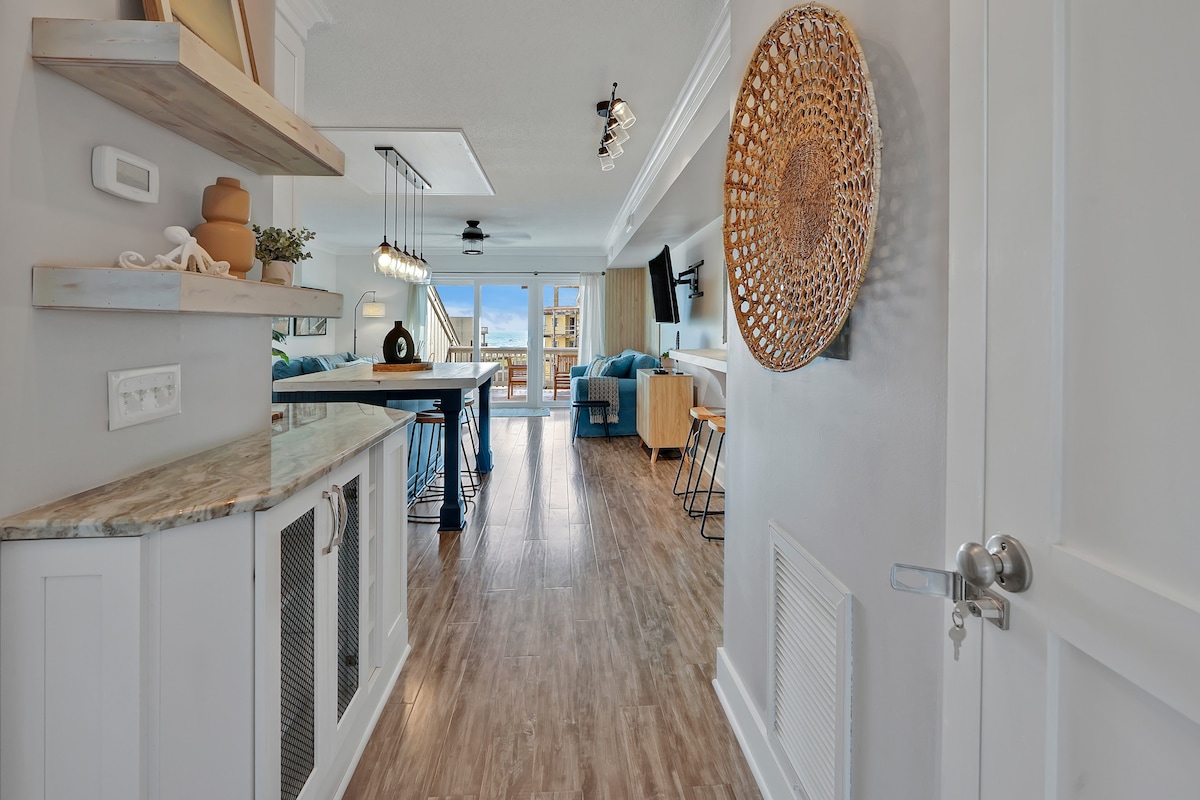 A modern entryway leads into a spacious open-concept living area. Natural light flows in through large windows, illuminating the wood-look tile flooring. The kitchen features textured granite countertops, while decorative shelving adds a coastal charm. A view of the balcony can be seen in the background.