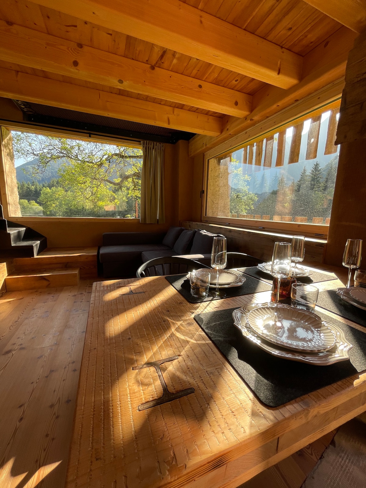 A dining area is positioned beside large windows that invite natural light and offer views of the surrounding greenery. A wooden table is set with glassware and decorative plates, complemented by a cozy seating area featuring a dark sofa near the entrance.