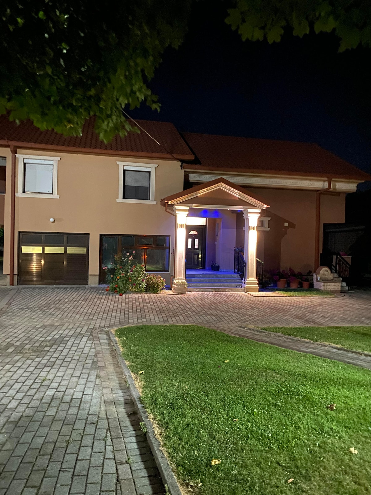The exterior of the villa is illuminated at night, featuring a grand entrance with a decorative canopy. Soft lighting highlights the architectural details, while a paved driveway leads to the entrance and is bordered by well-maintained grass and garden areas.