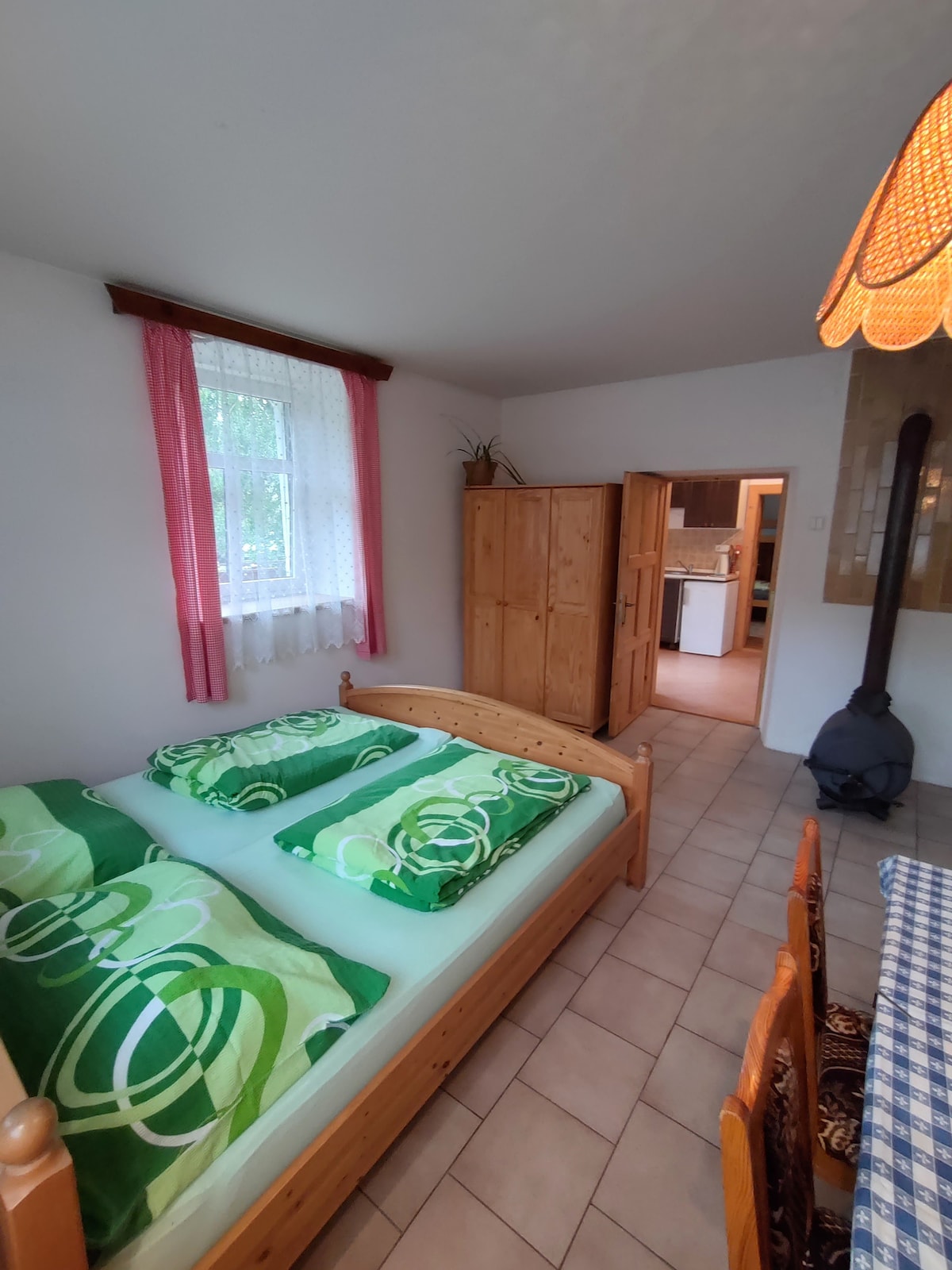 A cozy bedroom features a double bed with green bedding and two pillows. Natural light enters through a window adorned with sheer curtains. A wooden wardrobe and a dining area with a checkered tablecloth are visible in the background, enhancing the room's functionality.