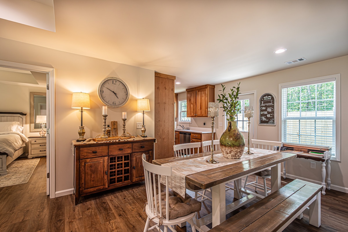 A dining area features a large wooden table surrounded by white chairs, creating a welcoming space for meals. Light-colored cabinets and a wall clock bring warmth to the area. Natural light filters through the windows, enhancing the inviting atmosphere.