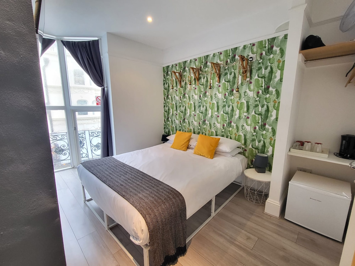 A bright room features a queen-sized bed accented with yellow pillows. The green wall art adds a lively touch, while a window with sheer curtains allows natural light to brighten the space. A small side table and a modern storage area are visible.