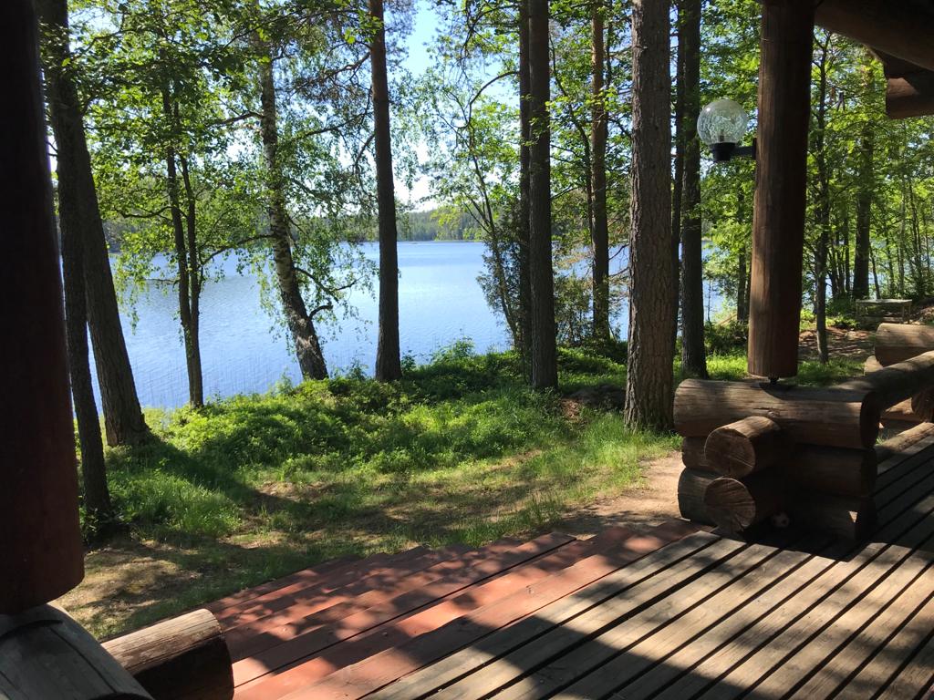 A view of a serene lake is framed by lush green trees from a wooden deck. The calm water reflects the surrounding nature, creating a peaceful atmosphere. The deck features log posts and smooth wooden flooring, offering a welcoming spot to relax.