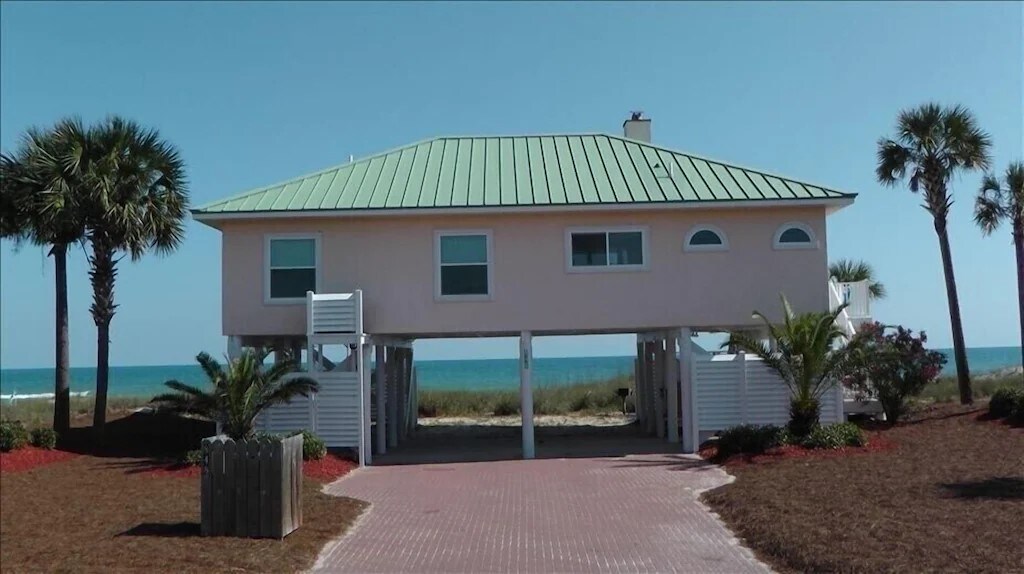 A beach house elevated on stilts, featuring a light pink exterior with a green metal roof. Palm trees are visible in the foreground, and the sandy beach can be seen in the background, leading to the ocean under a clear blue sky.