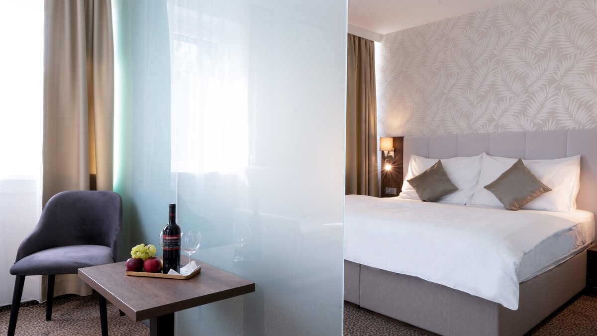 A modern bedroom features a spacious bed with neutral-toned linens and accented throw pillows. A small table with a bottle of wine, glasses, and fruit sits nearby. Natural light enters through a window, and soft curtains complement the decor.