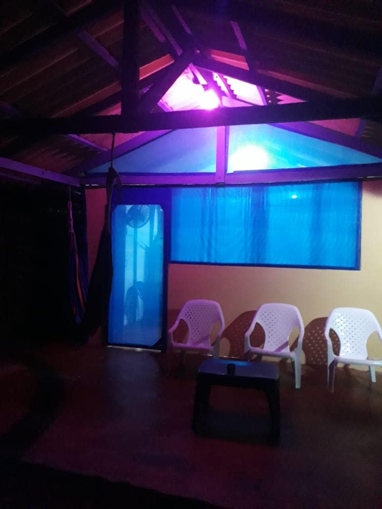 A spacious indoor area features a high, beamed ceiling with purple lighting and large windows adorned with blue curtains. Four white chairs and a small table are arranged, creating a comfortable setting for relaxation. A fan is positioned nearby, contributing to airflow.