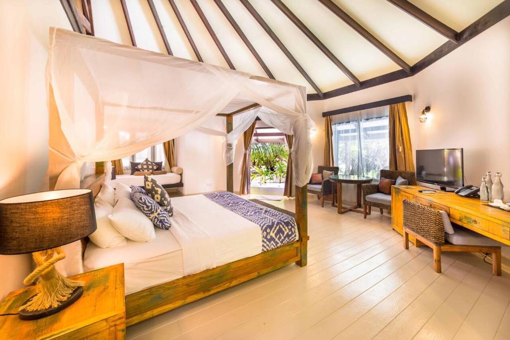 A spacious room is furnished with a canopied bed, accented by soft white linens and decorative pillows. Natural light filters through large windows, illuminating the wooden furniture, including a desk and lamp. A cozy seating area is visible, enhancing the layout's openness.