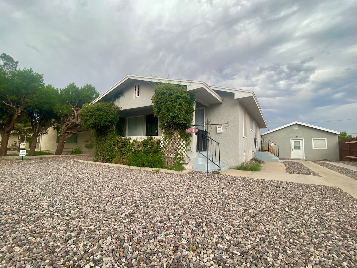 A one-story guest house is surrounded by laid stone and greenery. The exterior features a welcoming front entrance with steps leading up, flanked by compact shrubs and trees. A spacious driveway and an additional structure can be seen on the right.
