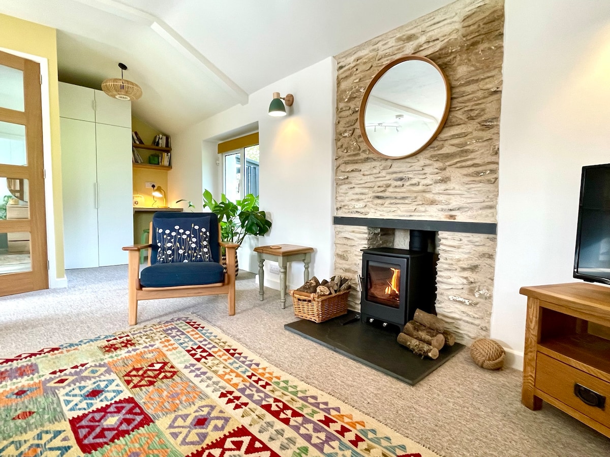A welcoming living space is highlighted by a wood-burning stove set against a textured stone wall. A round mirror hangs above the stove, and a colorful patterned rug is spread across the floor. Comfortable seating options include a blue armchair and a small table, with a glimpse of the workspace in the background.