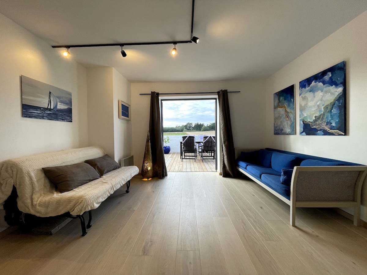 A spacious living area is presented with a light wood floor and two comfortable sofas. Large glass doors lead to an outdoor terrace, allowing natural light to fill the space. Artwork featuring coastal themes adorns the walls, enhancing the serene atmosphere.