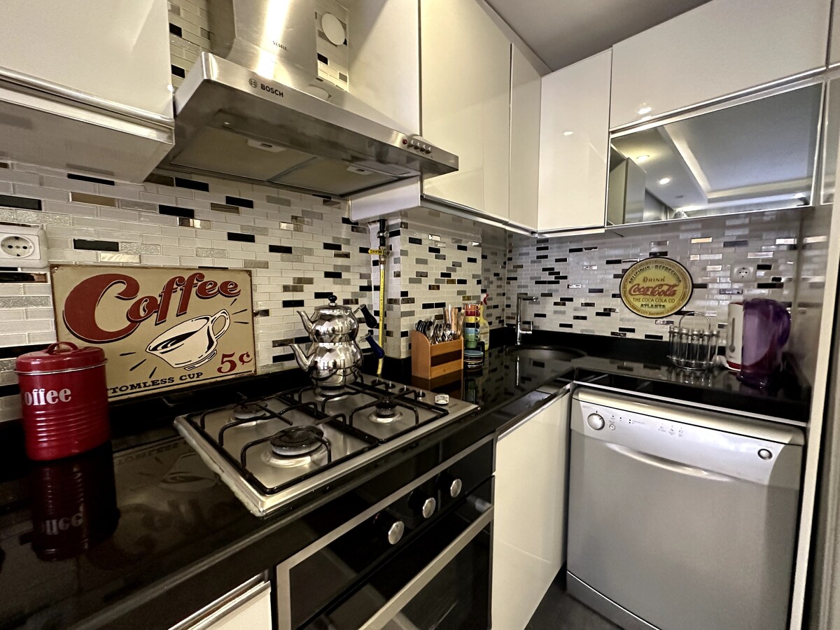 A modern kitchen features sleek black countertops and stylish cabinetry. Stainless steel appliances, including a gas stove and dishwasher, are visible. Decorative elements include a coffee-themed sign and a silver coffee pot, adding a homey touch to the contemporary design.