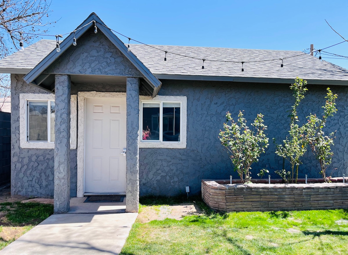 A charming exterior of a guest suite is shown, featuring textured grey walls and a welcoming white door. A window allows natural light into the space, while neatly landscaped greenery and flower beds are positioned in front, enhancing the overall appeal of the entrance.