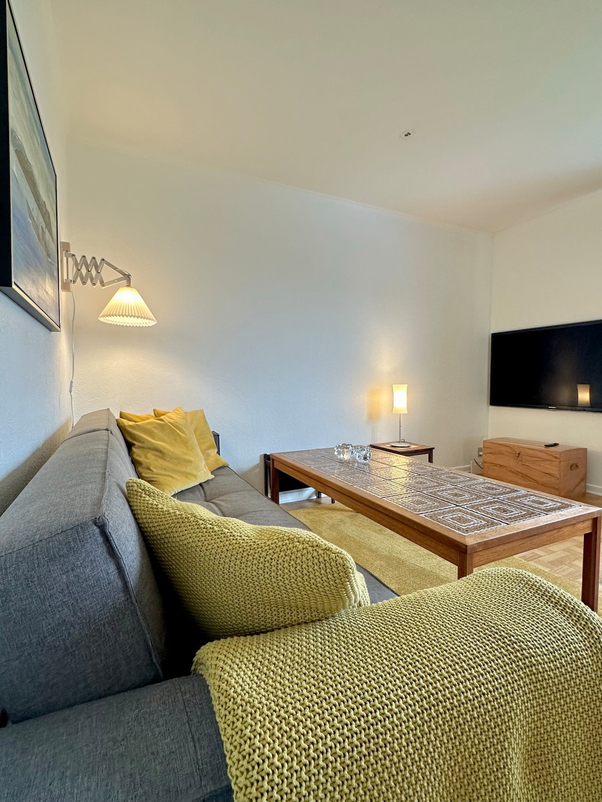 A cozy living area features a gray sofa with yellow accent pillows, complemented by a bright yellow knitted throw. A wooden coffee table holds decorative items and is positioned in front of a wall-mounted television. Soft ambient lighting is provided by lamps on the side table.