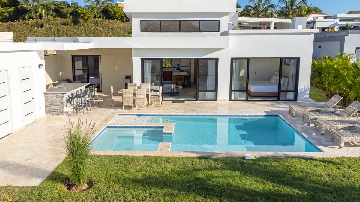 A modern villa is presented with a private pool surrounded by lounge chairs. An outdoor bar area complements the spacious patio. Floor-to-ceiling glass doors lead to the interior, where an inviting living space is visible. Lush greenery enhances the tranquil atmosphere.