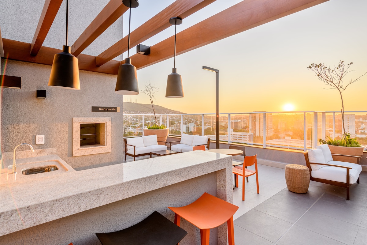 A rooftop space is visible, featuring a stone countertop with a grill, surrounded by modern seating arrangements in white and orange. The sunset casts a warm glow over the city skyline, enhancing the serene atmosphere.