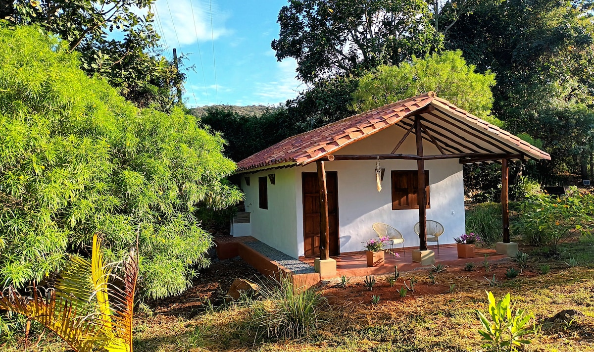 A charming cabin is framed by lush greenery and a well-maintained garden. The exterior showcases a natural aesthetic with a terracotta roof and wooden doors. A small porch is featured at the entrance, offering inviting seating among the tranquil surroundings.