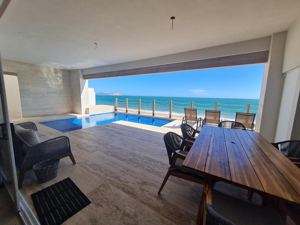 An outdoor seating area is paired with a large wooden dining table, offering views of the ocean. A swimming pool is visible, reflecting the clear blue sky, while the space is bordered by wooden railings, creating a seamless transition to the beach beyond.