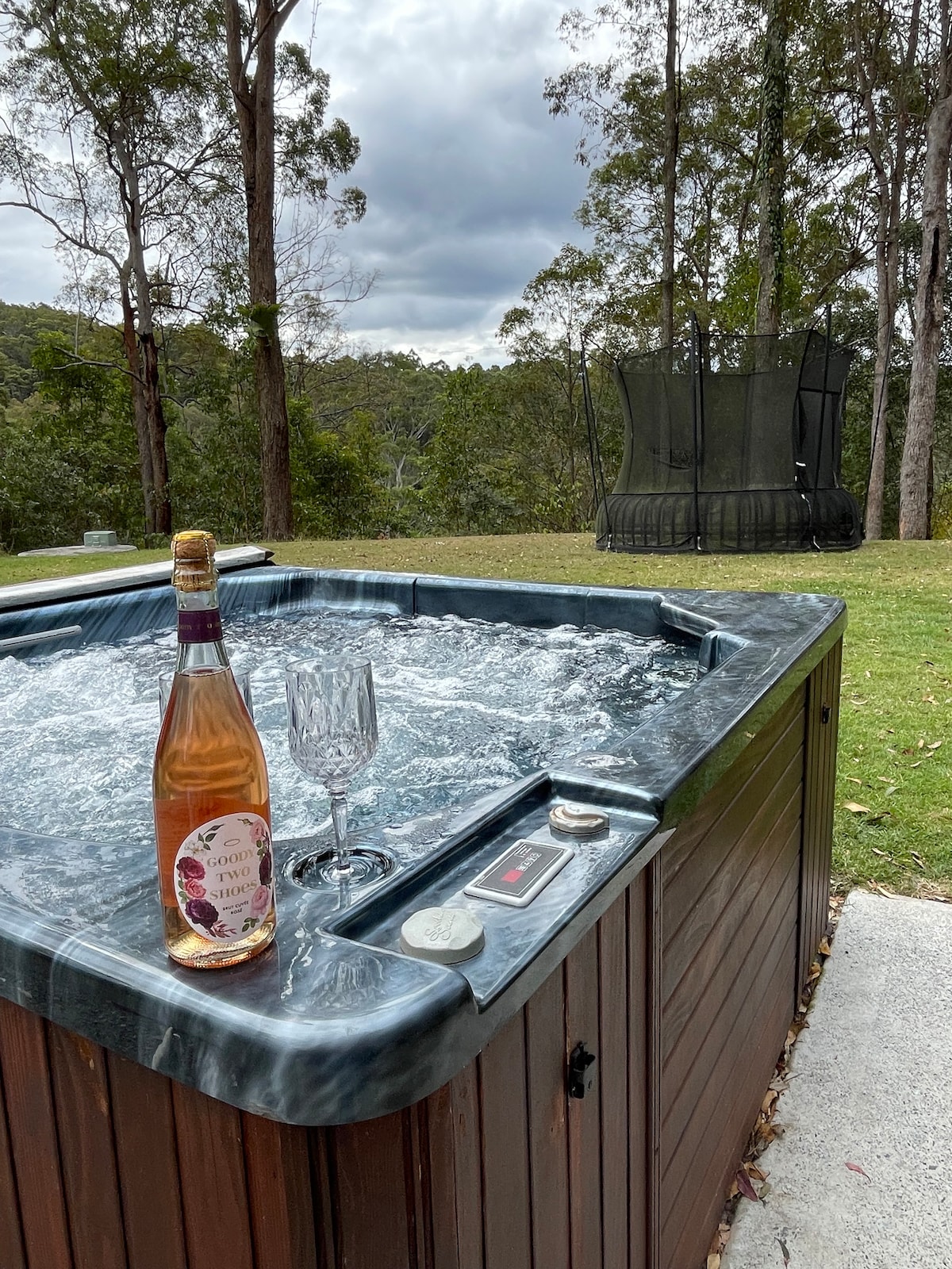 A relaxing hot tub is set amidst a natural backdrop, with bubbles gently rising to the surface. A bottle of wine and a glass rest on the edge, while a spacious green lawn and a trampoline are visible in the distance.