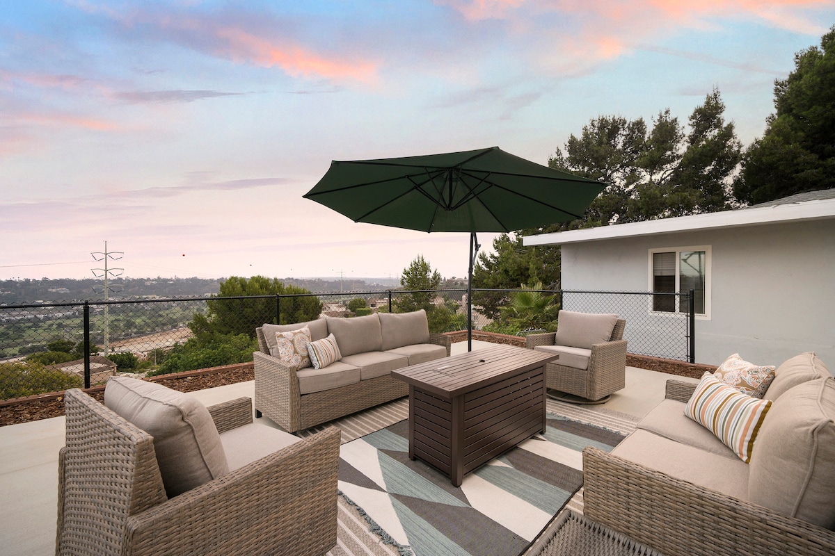 An outdoor seating area features a spacious arrangement of beige wicker furniture situated on a patterned rug. A large, green umbrella provides shade over a central coffee table. Panoramic views of the landscape and a pastel sky create a serene backdrop.