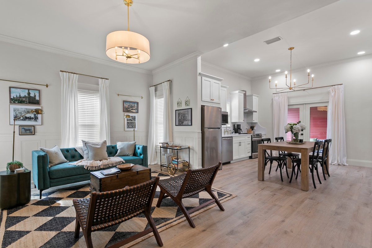 An open-concept living area features a plush teal sofa with assorted cushions. A dining table for four is positioned nearby, complemented by a modern chandelier and stylish chairs. Natural light enhances the space, reflecting off the hardwood floor and highlighting the kitchen area with sleek appliances.