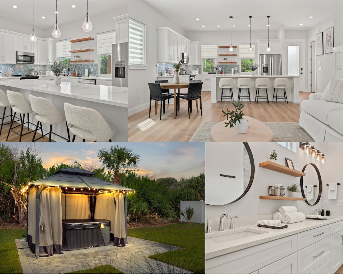 The image displays a modern kitchen with white cabinetry and stainless steel appliances. A dining table with seating for four is positioned beside a kitchen island with additional seating. An outdoor area features a hot tub surrounded by a patio, elegantly lit by string lights.