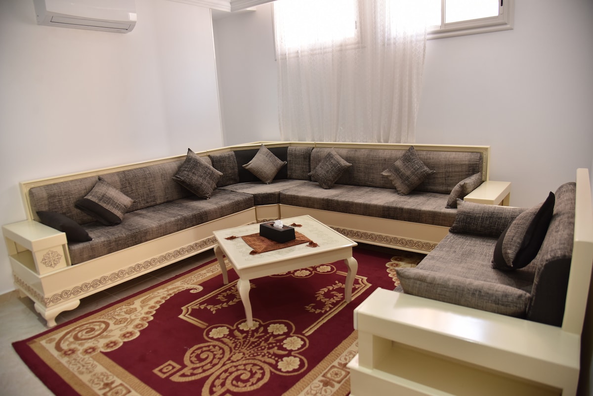 A comfortable seating area is featured, with a large sectional sofa adorned with plush cushions. A coffee table sits in the center, placed on a decorative red area rug. Natural light enters through the windows, brightening the modern, minimalist décor.