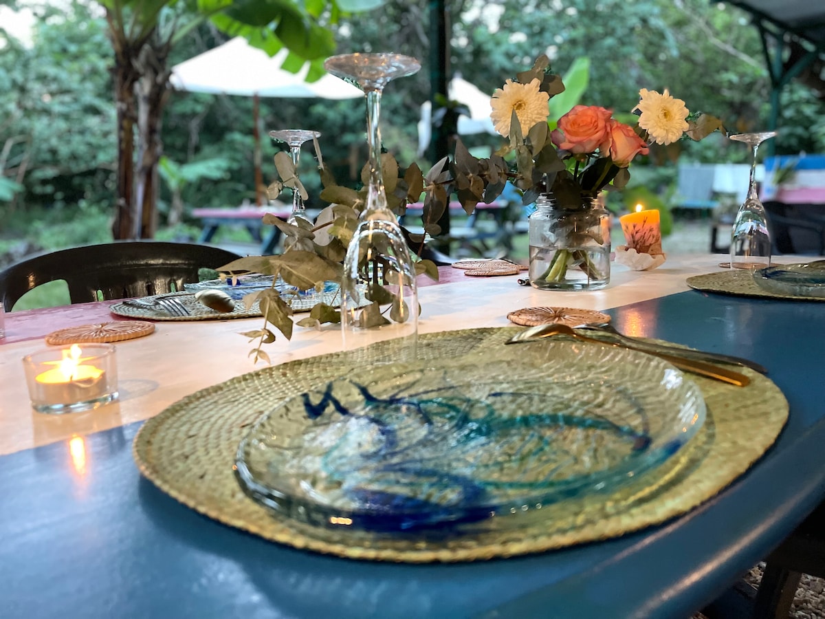 A dining table is set outdoors with natural greenery in the background. Clear glasses and elegant plates are placed on woven placemats. A centerpiece of fresh flowers adds a touch of color. Candles provide soft lighting, enhancing the serene atmosphere.