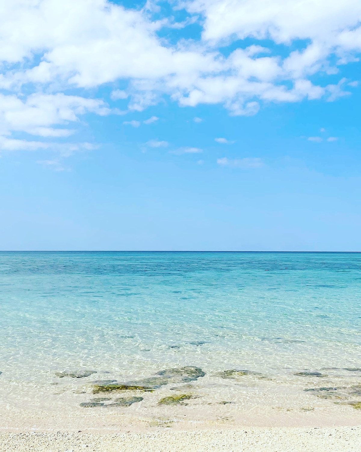 A serene beach scene presents clear turquoise waters gently lapping against a sandy shore. Soft clouds drift across a bright blue sky, while rocky formations are visible beneath the water's surface, creating a tranquil and inviting atmosphere for relaxation and exploration.