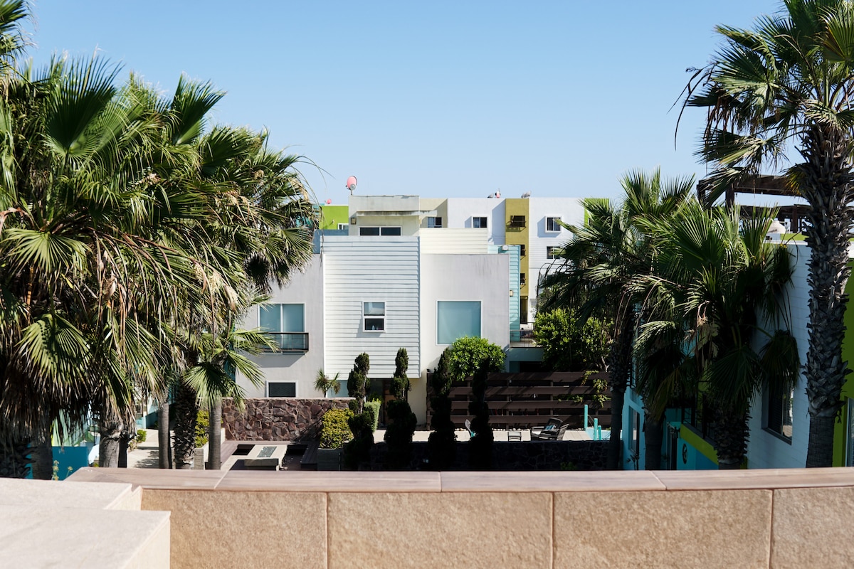 A view from a balcony showcases a landscaped courtyard surrounded by palm trees. Colorful residential buildings are visible in the background, creating a vibrant atmosphere. Clear blue skies complement the serene setting.