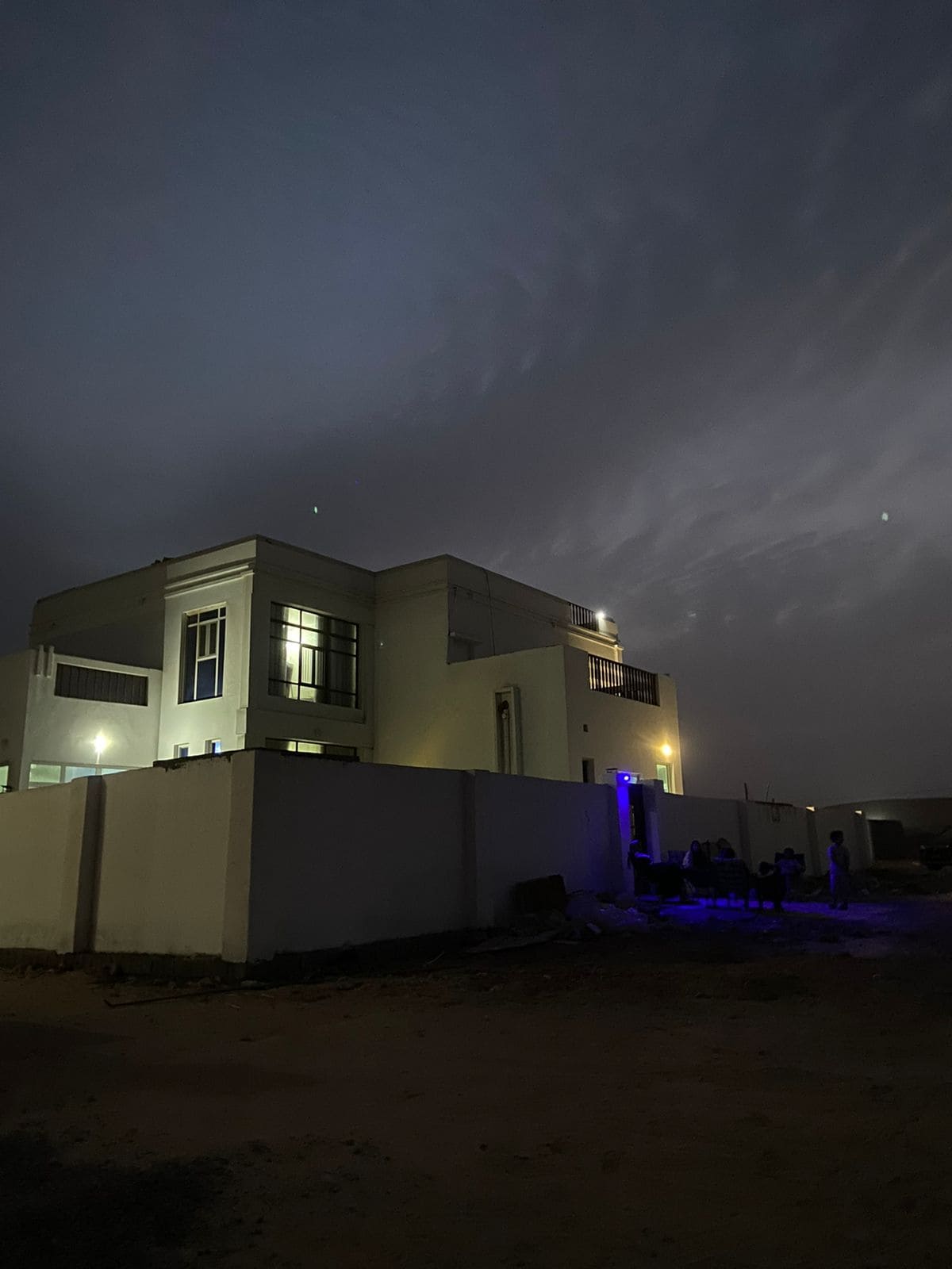 A modern two-story villa is featured at night, illuminated by soft outdoor lighting. The facade showcases large windows, and the structure is surrounded by a low wall. A clear sky with faint clouds is visible above, creating a serene atmosphere.