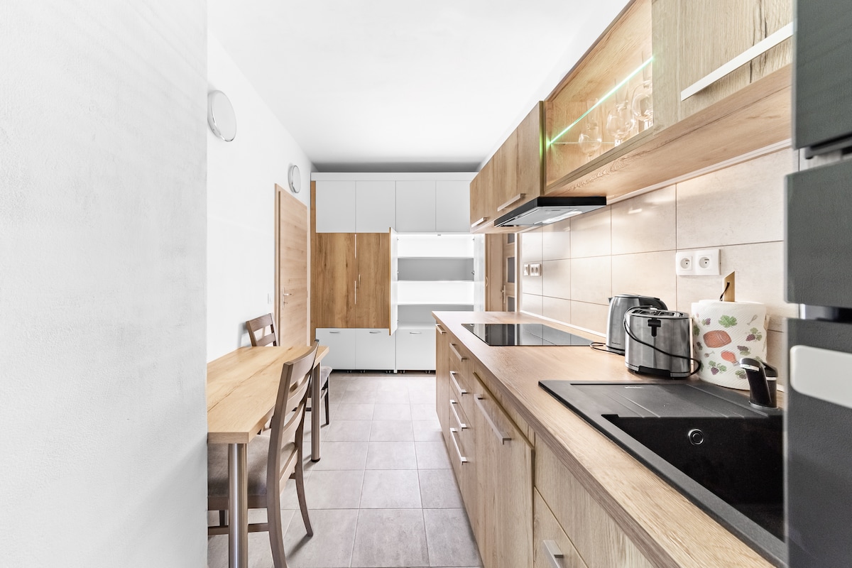 A modern kitchen features light wood cabinetry and a spacious countertop. Equipped with essential appliances, it includes a dining table with seating for four. Soft lighting illuminates the area, highlighting the sleek design and tile flooring.