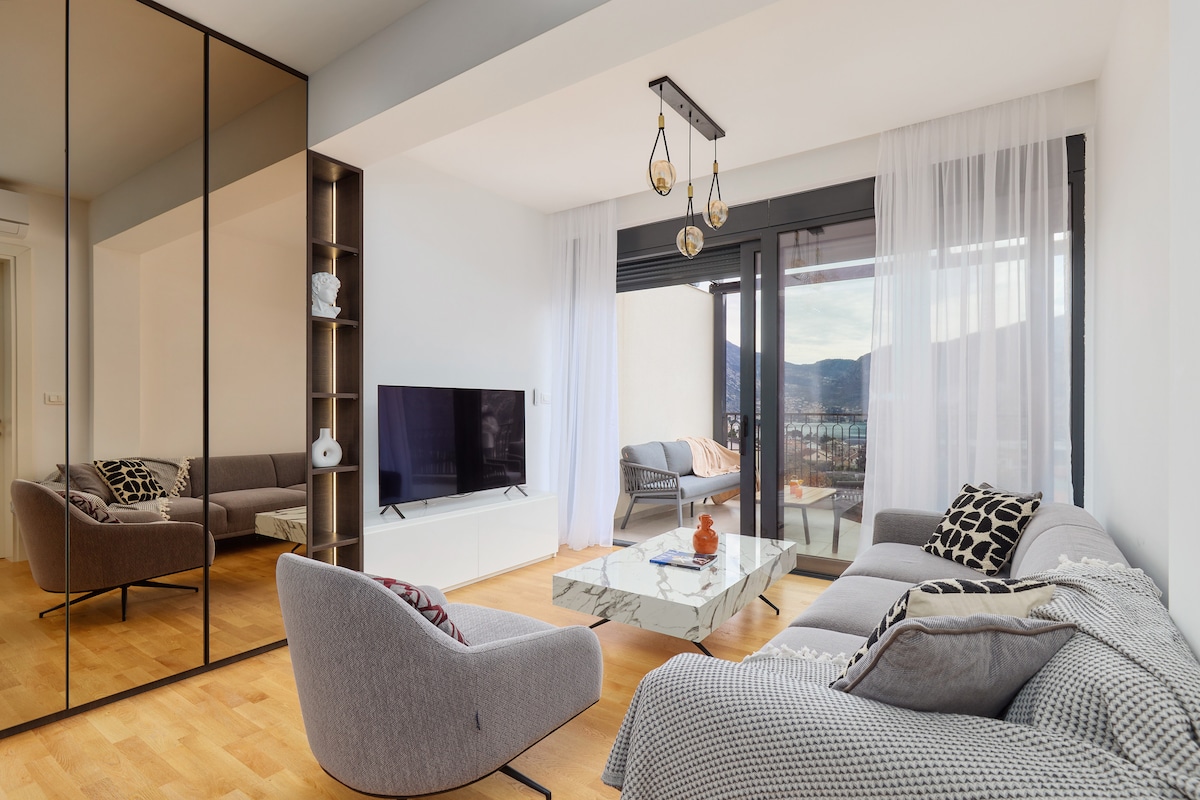 A contemporary living area features large windows that allow natural light to fill the space. Two comfortable sofas, a marble coffee table, and a mounted television create a cozy setting. Decorative accents, such as pillows and a plant, enhance the environment's modern appeal.