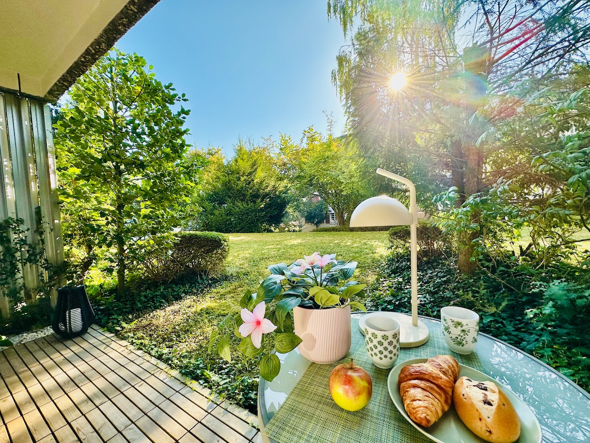 A serene terrace is presented, featuring a small round table set with a plant, two cups, an apple, and croissants. Lush greenery surrounds the view, while sunlight filters through the trees, creating a peaceful outdoor dining space.