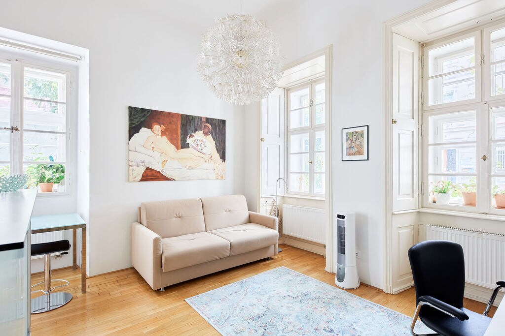 A bright living area features large windows adorned with potted plants, allowing natural light to fill the space. A neutral-toned sofa sits opposite a light wood table, and an artistic wall hanging adds a touch of character. A modern chandelier illuminates the room.