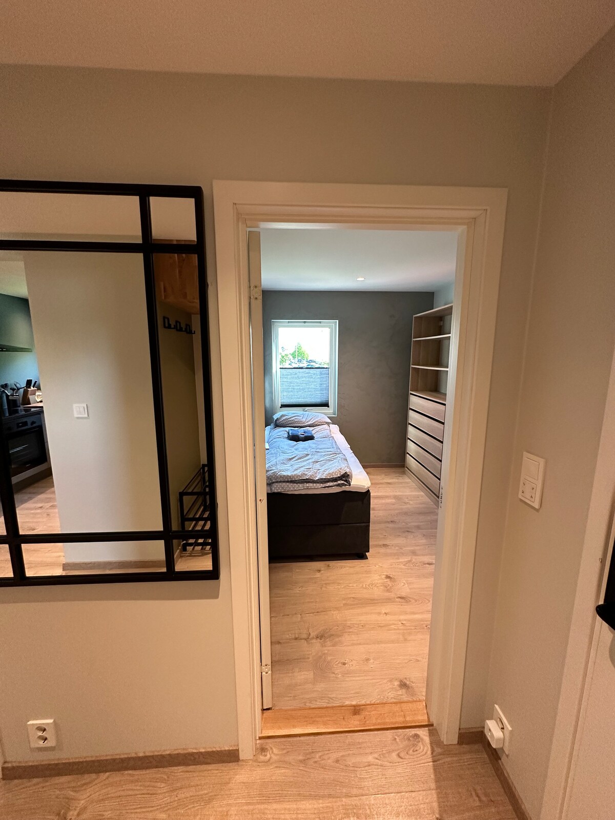 A doorway leads into a well-lit room featuring a spacious bed with a light blanket. The neutral-colored walls complement a simple wooden dresser against the opposite wall. A window allows natural light to enter, enhancing the inviting atmosphere of the space.