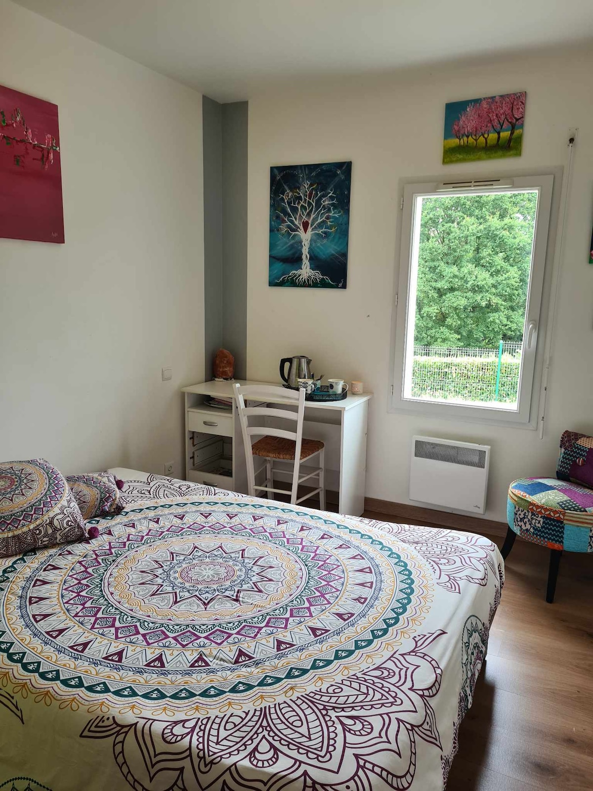 A comfortable sleeping area is highlighted by a colorful patterned bedspread covering a spacious bed. A desk with a kettle and cups is positioned beside the window, allowing natural light to illuminate the space. A chair with vibrant upholstery adds a splash of color to the room.