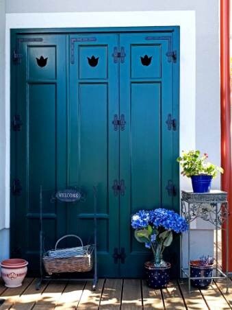A dark green double door is framed with decorative iron hardware, featuring cut-out shapes at the top. A small table stands beside the door, adorned with a potted plant and blue flowers. A woven basket rests on the ground, enhancing the welcoming entryway.