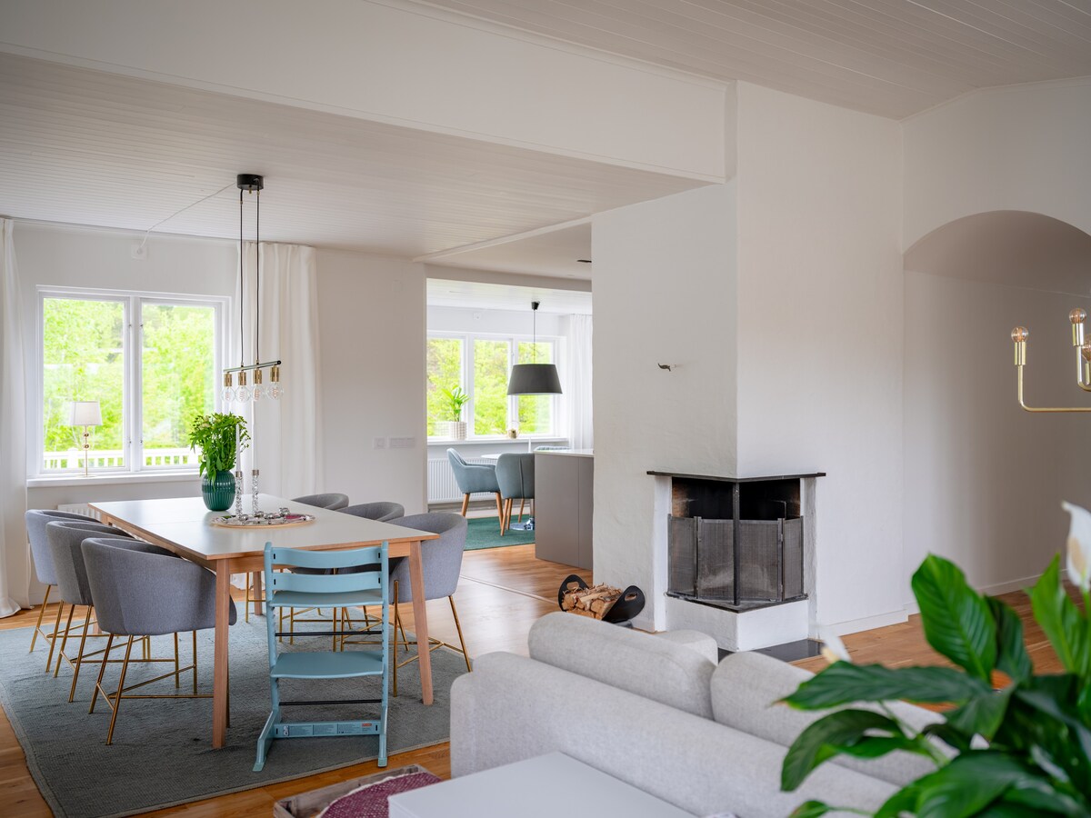 An open-plan living and dining area is illuminated by natural light pouring in through large windows. A wooden dining table surrounded by chairs sits adjacent to a cozy sitting area with a neutral-colored sofa. A modern wood-burning stove adds warmth to the space.