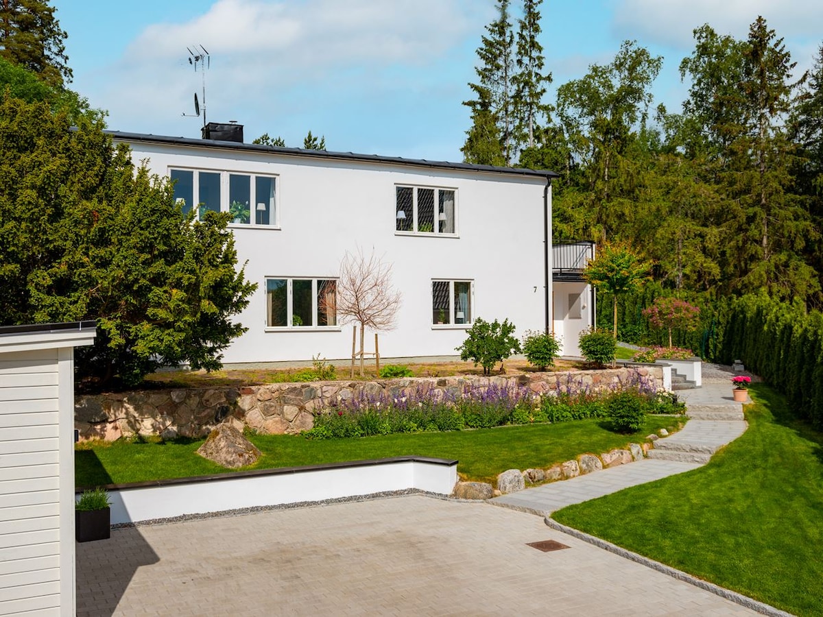 A modern two-story villa is set within a landscaped area, featuring a manicured lawn and colorful flower beds. The exterior is white with large windows, allowing for ample natural light. Pathways lead to the entrance amidst trees and greenery.