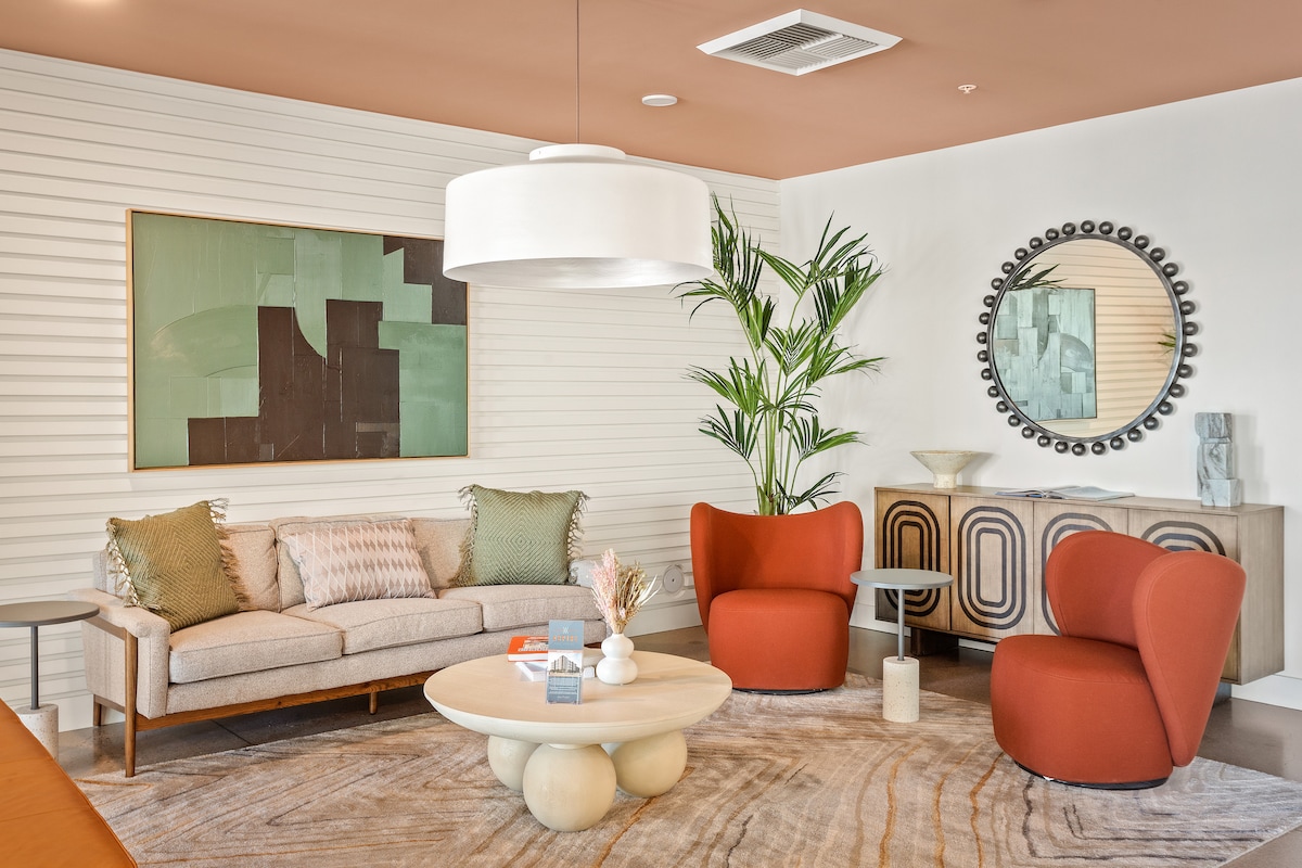 A modern living area is highlighted by a light-colored sofa with decorative pillows and two red chairs. A round coffee table sits in the center on a textured area rug, complemented by a large pendant light and a decorative mirror on the wall.