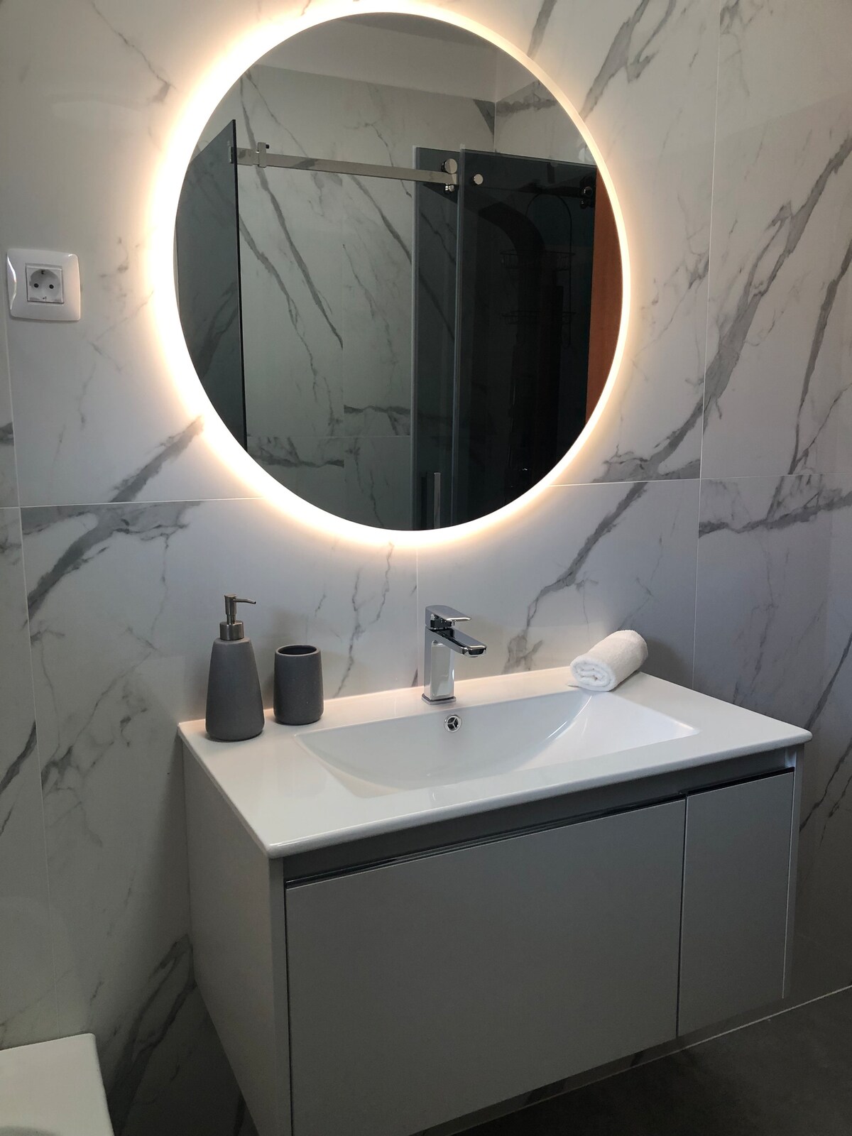 A modern bathroom is presented with sleek, marble-patterned walls. A round mirror with backlighting is mounted above a minimalistic sink. The vanity unit features a smooth surface and a simple design, complemented by two decorative bottles and a rolled towel.