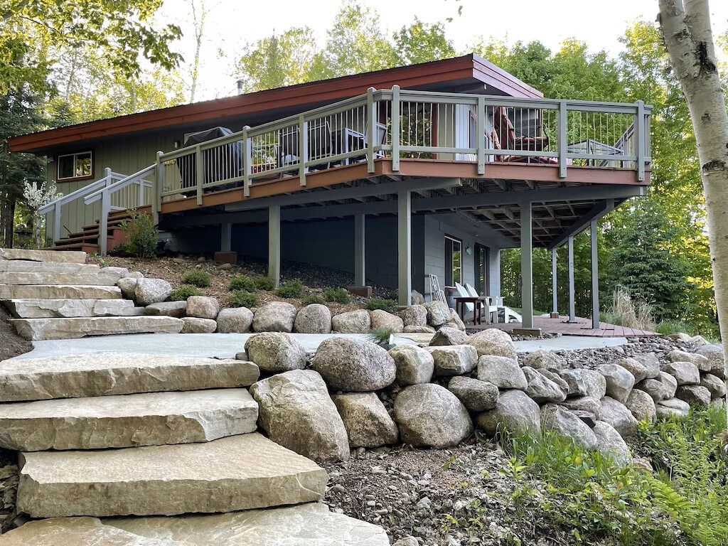 A modern home sits elevated on a landscaped property, featuring a spacious deck surrounded by greenery. Stone steps lead up to the entrance, highlighting the home’s outdoor charm and accessibility. Natural light filters through the trees, creating a serene atmosphere.