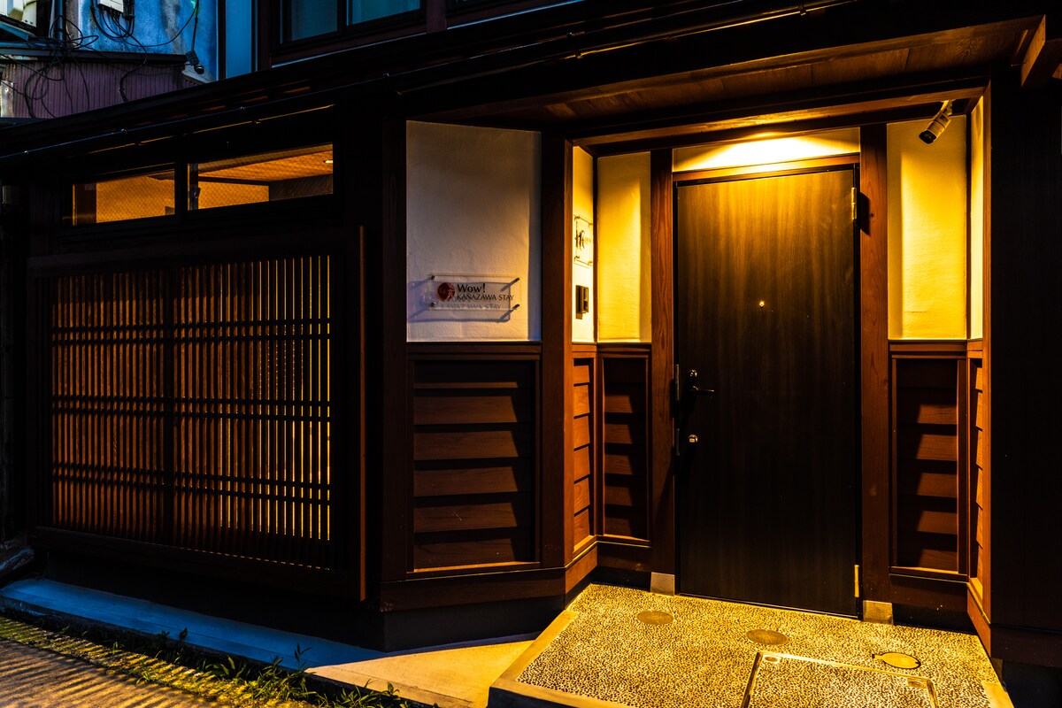 A traditional entrance to the guesthouse features a dark wooden door framed by warm lighting. The façade includes wooden slats, creating a subtle pattern. A solid stone pathway leads to the door, enhancing the welcoming ambiance of the location.