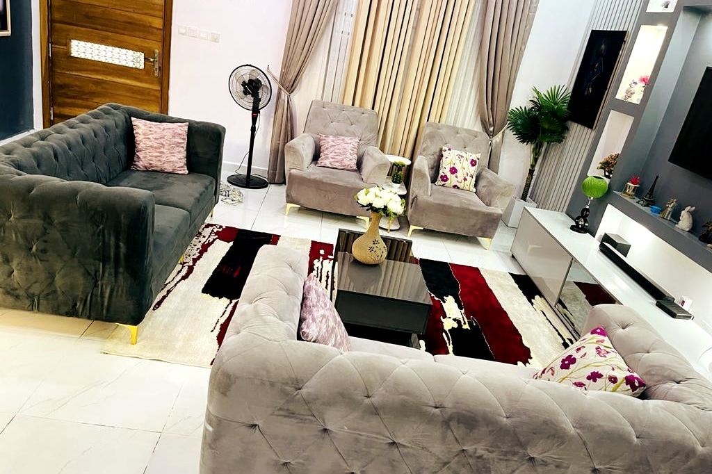 A comfortable seating area features gray upholstered sofas and armchairs, arranged around a modern coffee table. A decorative rug with bold patterns anchors the space. Soft curtains frame large windows, allowing natural light to enter, while plants add a touch of greenery.